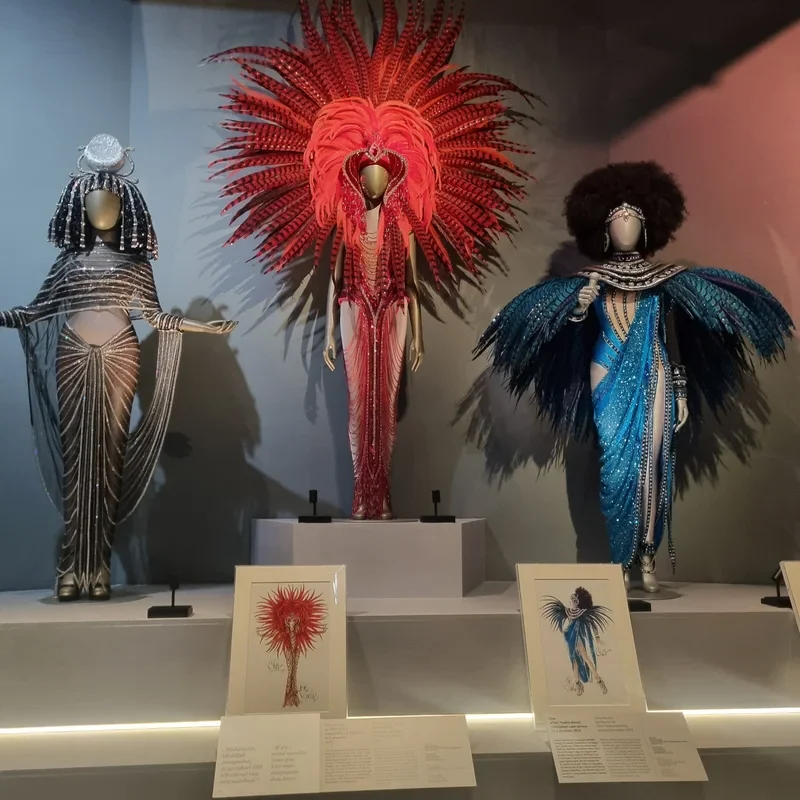Three ornate mannequined costumes with dramatic feather headdresses in red, blue, and silver, displayed on a gallery wall with info cards nearby.