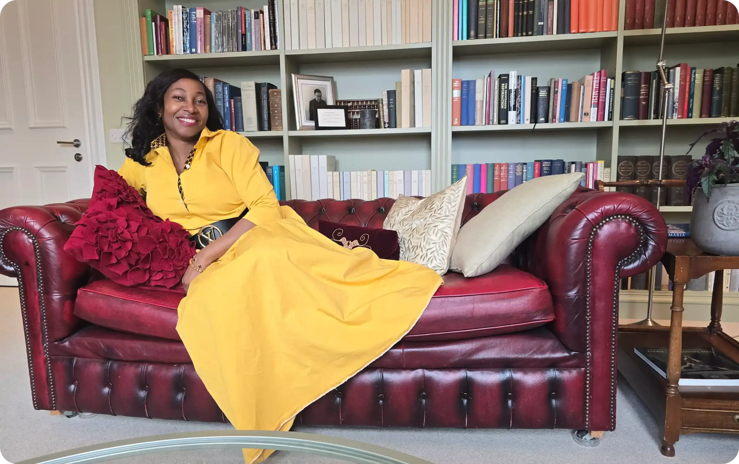 Smiling woman in a bright yellow dress relaxing on a red leather sofa with bookshelves in the background.