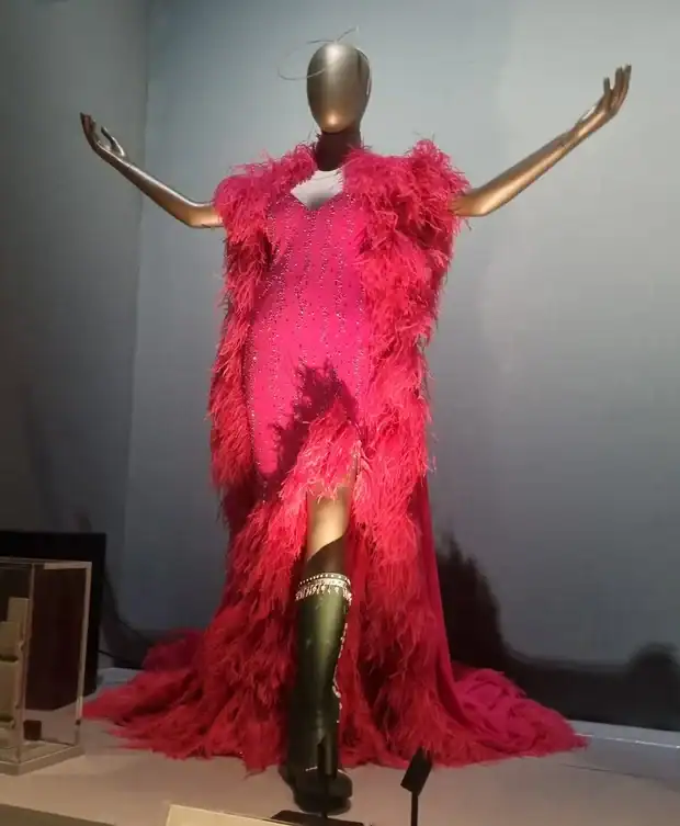 Mannequin in a bright pink feathered evening gown with a sparkling bodice, posed with arms outstretched in a gallery display.