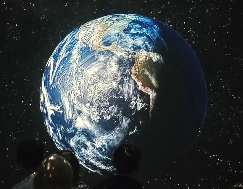 20260702_161327 Earth as seen from space with continents and swirling clouds, viewed by silhouettes of visitors in a dark room.