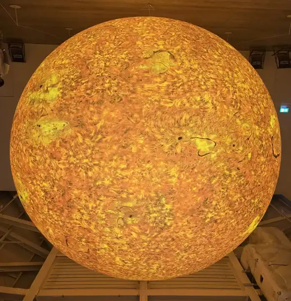 8 Giant orange-yellow sphere on display indoors, resembling the Sun, in a museum or science center setting.